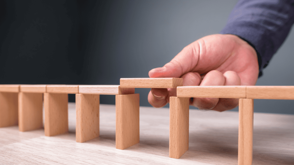 A person's hand placing a wooden block atop a neatly aligned row of small wooden blocks, symbolizing the importance of a business website in forming a structured foundation on a light wooden table against a soft gray background.