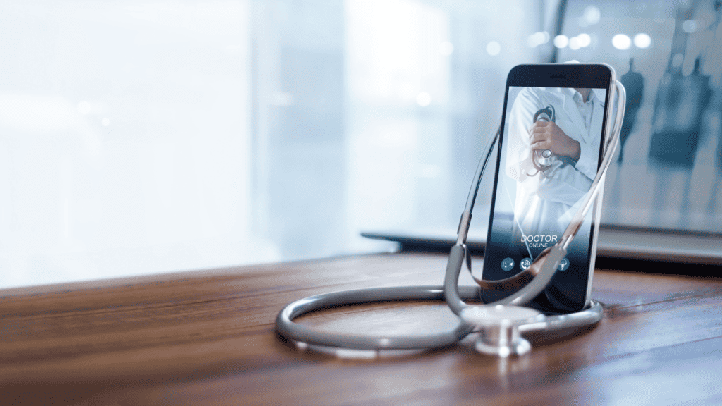 A smartphone on a wooden table displaying a video call with a doctor, who is visible on screen holding a stethoscope. A stethoscope also lies on the table in front of the phone, emphasizing remote medical consultation and showcasing social media marketing strategies for hospitals.