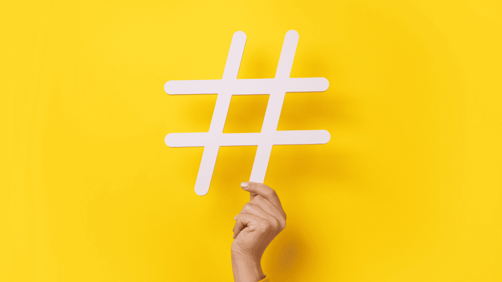 A hand holding a large white hashtag symbol against a bright yellow background. The fingers grasp the base of the vertically positioned symbol, emphasizing its outline and contrast, suggesting innovative social media marketing strategies for hospitals.