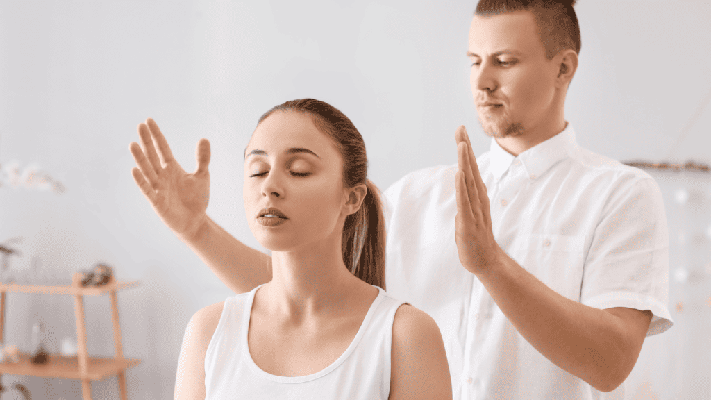 A man performs a reiki healing session on a woman, both dressed in white. They stand indoors with hands raised but not touching, eyes closed, focusing on therapy. Soft, warm background with decor enhances the therapeutic atmosphere ideal for promoting wellness-focused clothing brands on social media.