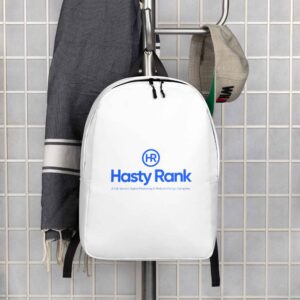 A white HR Urban Voyager Backpack with the "hasty rank" logo hangs on a hook in a tiled wall, alongside a grey jacket and a cap. The backpack is prominently displayed against the neutral backdrop.