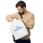 A man with a beard examines a white HR Urban Voyager Backpack that he is holding. the backpack features the "hasty rank" logo in blue lettering inside a blue circle. he wears a beige long-sleeve shirt and a black shoulder strap crosses his torso.