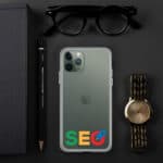 Top view of a desk with a green SEO Clear Case for iPhone® labeled "seo" on google's logo colors, a gold watch, glasses, a notebook, and a pencil, all organized neatly on a black surface.