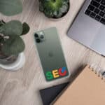 An SEO Clear Case for iPhone® with a green case displaying the "seo" logo in colorful letters is placed on a wooden desk beside a laptop, a notebook, a small potted plant, and a speaker.