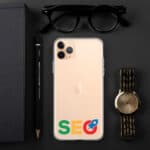 Top view of a desk with a gold SEO Clear Case for iPhone® featuring a google seo sticker, a golden watch, black reading glasses, a book, and a pen, all arranged neatly on a dark surface.