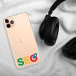 A smartphone with a gold back, displaying the letters "seo" and a google logo, lies on a plush white fabric. SEO Clear Case for iPhone® with a strap rest partially draped over the phone.