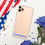 An iPhone with a HR CrystalShield iPhone® Case placed on a surface with an American flag partially visible, scattered red confetti, and blue flowers. The phone displays a generic company logo for "hasty rank" on its case.