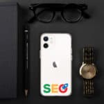 Flat lay of a workspace with a white SEO Clear Case for iPhone®, a black notebook, a gold wristwatch, a pencil, and sunglasses on a dark background.