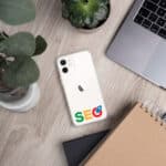 A white smartphone with an SEO Clear Case for iPhone® case and Google logo sticker on the back, lying next to a laptop and a notebook on a wooden desk, surrounded by a small green plant and a potted succulent.
