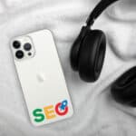 A white smartphone with an "SEO Clear Case for iPhone®" sticker and google logo, lying on a soft white blanket, next to black over-ear headphones. the phone display faces up, showing the apple logo and three rear cameras.