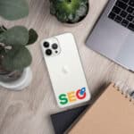 A white smartphone with a triple camera setup lies on a wooden desk, adorned with colorful SEO Clear Case for iPhone® stickers. nearby are a laptop, a notebook, a small potted plant, and a speaker.
