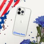 A silver HR CrystalShield iPhone® Case with a triple camera setup on a white background, partially covered by an american flag and scattered red and blue confetti, next to blue flowers. a sticker labeled "hasty rank" is on the phone.
