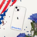 A smartphone with a HR CrystalShield iPhone® Case showcasing the logo "hasty rank" at the bottom back, placed on a flat surface with an american flag and blue flowers scattered with small red and blue stars.