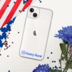 A HR CrystalShield iPhone® Case with a blue "hasty rank" logo on the bottom covers an iphone with a dual-lens camera. it lies on a surface adorned with a u.s. flag and blue flowers, scattered with small red and blue stars.