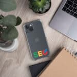 A smartphone with a SEO Clear Case for iPhone® displaying the word "seo" in colorful letters on a desk next to a laptop, notebook, and potted plant. The office setup suggests a focus on digital marketing or web optimization.