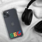 A SEO Clear Case for iPhone® with an "seo" logo on a gray iphone, resting on a soft white blanket next to black over-the-ear headphones. the case highlights the apple logo and the phone's camera layout.