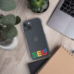 A SEO Clear Case for iPhone® with a google logo on the case lying on a wooden desk next to a laptop, a notebook, and a potted plant, showing a modern workspace setting.