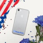 A HR CrystalShield iPhone® Case with a visible apple logo is centered on a white surface, partially covered by a draped american flag and scattered small red and blue stars, with blue flowers at the corner.
