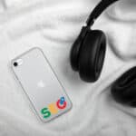 A smartphone with a SEO Clear Case for iPhone®, displaying a modified google logo reading "seo," lies on a white fluffy blanket next to a black over-ear headphone. the environment suggests cozy and relaxed tech use.