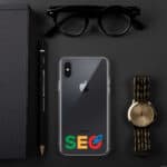 Flat lay of a workspace featuring an SEO Clear Case for iPhone®, a gold wristwatch, black glasses, a black notebook, and a pen, all arranged on a dark surface.
