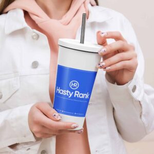 A person wearing a pink hoodie and a white jacket holds an HR SplashGuard Tumbler with a blue label featuring "hasty rank" logo. they have manicured nails with artistic patterns, using a metal straw to drink.