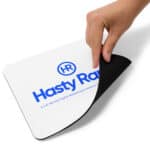 A hand is partially lifting a corner of a rectangular HR PrecisionPad Mouse Pad displaying "hasty rank, hr" logo in blue, with text "a full-service digital marketing & website co." on a white background.