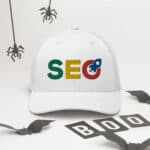 A white SEO Professional Cap with "seo" in colorful letters on the front, surrounded by halloween decorations including hanging spiders and bat cutouts, with the word "boo" visible on the ground.
