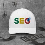 A white SEO Professional Cap with the letters "seo" in green, yellow, and red, featuring a magnifying glass on the letter 'o'. the cap is on a grey textured background with hanging spiders and paper cutouts that spell "boo".