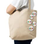 A person from the side wearing a plain beige canvas tote bag with six white circular badges, each featuring the letters "seo" in colorful, overlapping text. the background is white and unadorned.