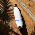 A white HR Elite Hydration Flask with "hasty raj" in blue text, lying on a rustic wooden surface dappled with tree shadow patterns, highlighted by sunlight.