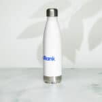 A white insulated stainless steel HR Elite Hydration Flask with a metallic cap and base, featuring a blue "vrank" logo, stands on a light gray surface with soft shadow patterns of leaves on the wall behind it.