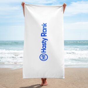 A person stands on a sandy beach holding up a large white HR Signature Towel with its blue logo. The background shows a clear sky and calm sea.