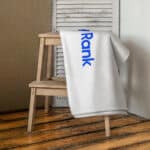 A white HR Signature Towel with the word "rank" in blue letters draped over a wooden chair against a rustic wooden floor and a grey shuttered window background.