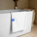 A white HR Signature Towel with the word "think" in blue letters draped over the side of a modern, rectangular bathtub in a beige-tiled bathroom. decorative dried plants are in the background.