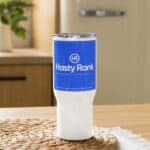 A promotional HR Journey Companion Mug is displayed on a wicker placemat on a wooden table. The mug is white at the base and transitions to blue at the top with the logo "hr hasty rank" and additional text about the company.