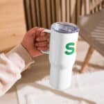 A person's hand holding a white insulated "SEO Travel Mug" with a green letter "s" printed on it. the mug has a clear, sliding lid. the background depicts a soft-focus wooden floor and part of a chair.