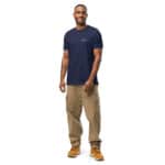 A smiling man stands confidently, wearing a navy blue HR PrimeStyle Tee with the text "go beyond" and beige cargo pants, complemented by tan boots. he has a visible tattoo on his left arm.