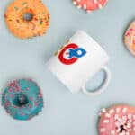 A SEO White Glossy Mug with a colorful logo sits centered among various decorated doughnuts on a light blue background. the doughnuts feature sprinkles, and confections in shades of orange, pink, and blue.