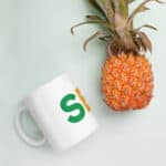 A SEO White Glossy Mug with a green letter 's' on it rests near a ripe pineapple on a light green background. the image is shot from above, displaying a clean and simple composition.