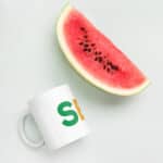 A slice of juicy watermelon with black seeds beside a SEO White Glossy Mug with a green and orange "s" logo, both placed on a light gray background.