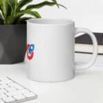 A SEO White Glossy Mug with a colorful logo featuring a red speech bubble and a blue ear, placed on a desk next to a keyboard and a green plant, with a white notebook in the background.